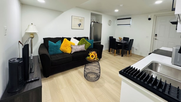 A modern living room with a black sofa, a coffee table with a vase of sunflowers, a dining area with a table and chairs, and a kitchen with a sink and stove.