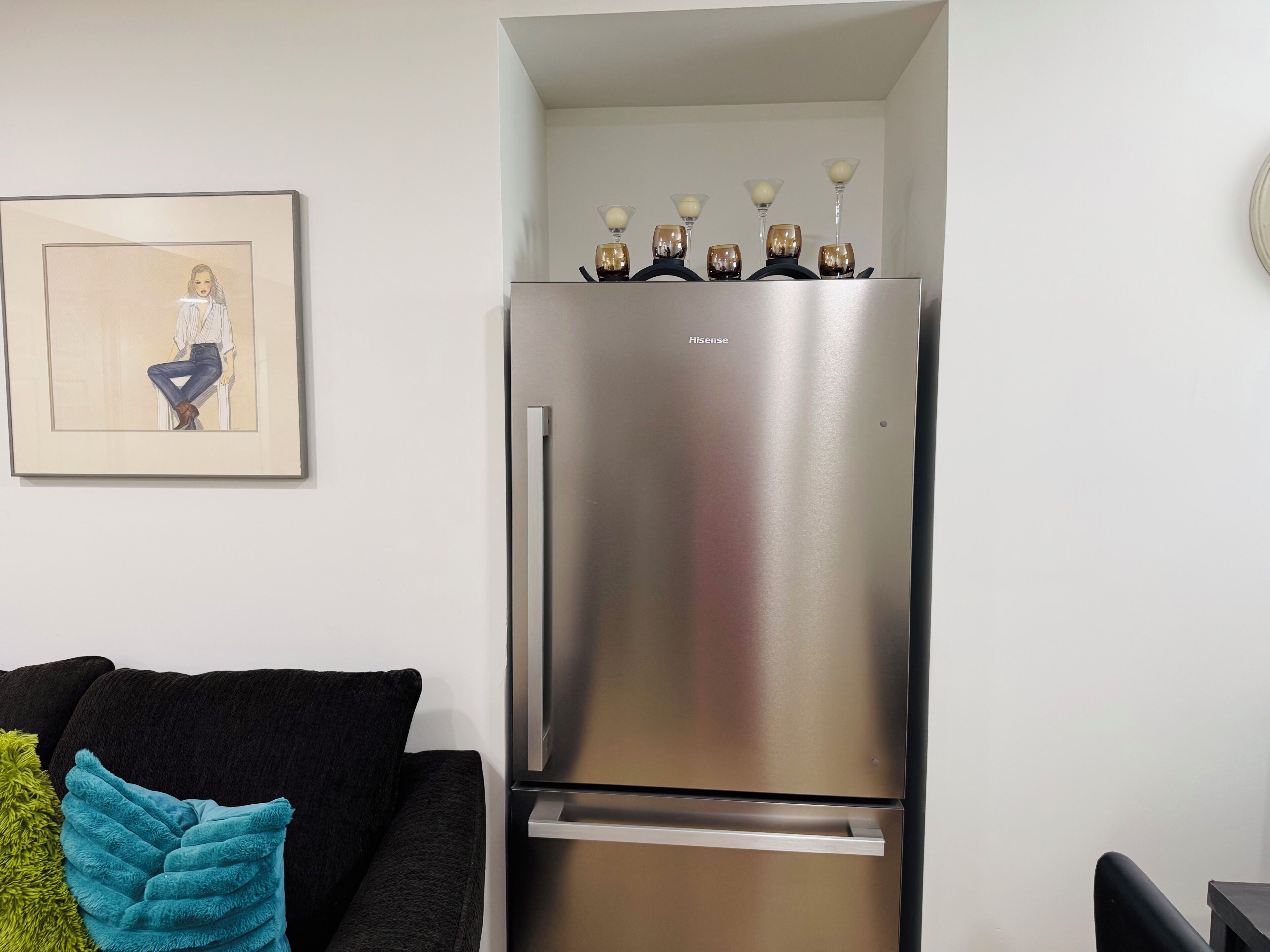 A modern kitchen with a stainless steel refrigerator, a framed artwork on the wall, and a black sofa with a teal throw pillow.