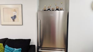 A modern kitchen with a stainless steel refrigerator, a framed artwork on the wall, and a black sofa with a teal throw pillow.