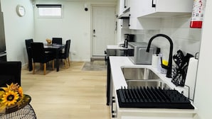 A modern kitchen with white cabinets, a stainless steel sink, and a black faucet. There is a dining area with a table and chairs, and a small decorative plant with sunflowers.