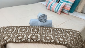 A bed with a beige duvet, a brown and white crocheted blanket, and a rolled-up grey towel.
