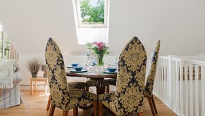 A dining area with a table set for four, chairs with ornate patterns, a window with a view of greenery, and a white railing along the wall. - The Coach House - Beautiful 3 Bedroom Home in Oxfordshire (Souldern)