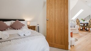 A bedroom with a bed, pillows, a bedside table, a lamp, and a wooden door leading to a dining area with a table and chairs. - The Coach House - Beautiful 3 Bedroom Home in Oxfordshire (Souldern)