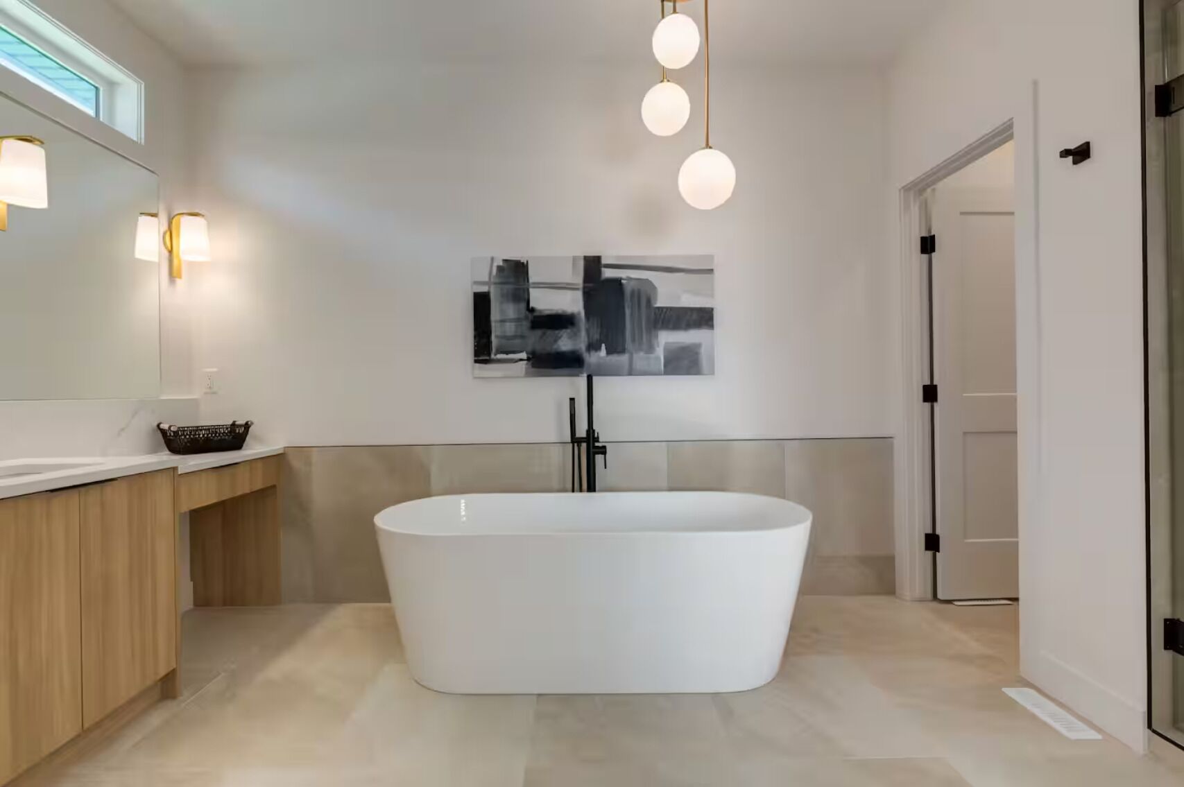 A modern bathroom with a freestanding bathtub, a wooden vanity, a wall-mounted light fixture, and a framed abstract painting.