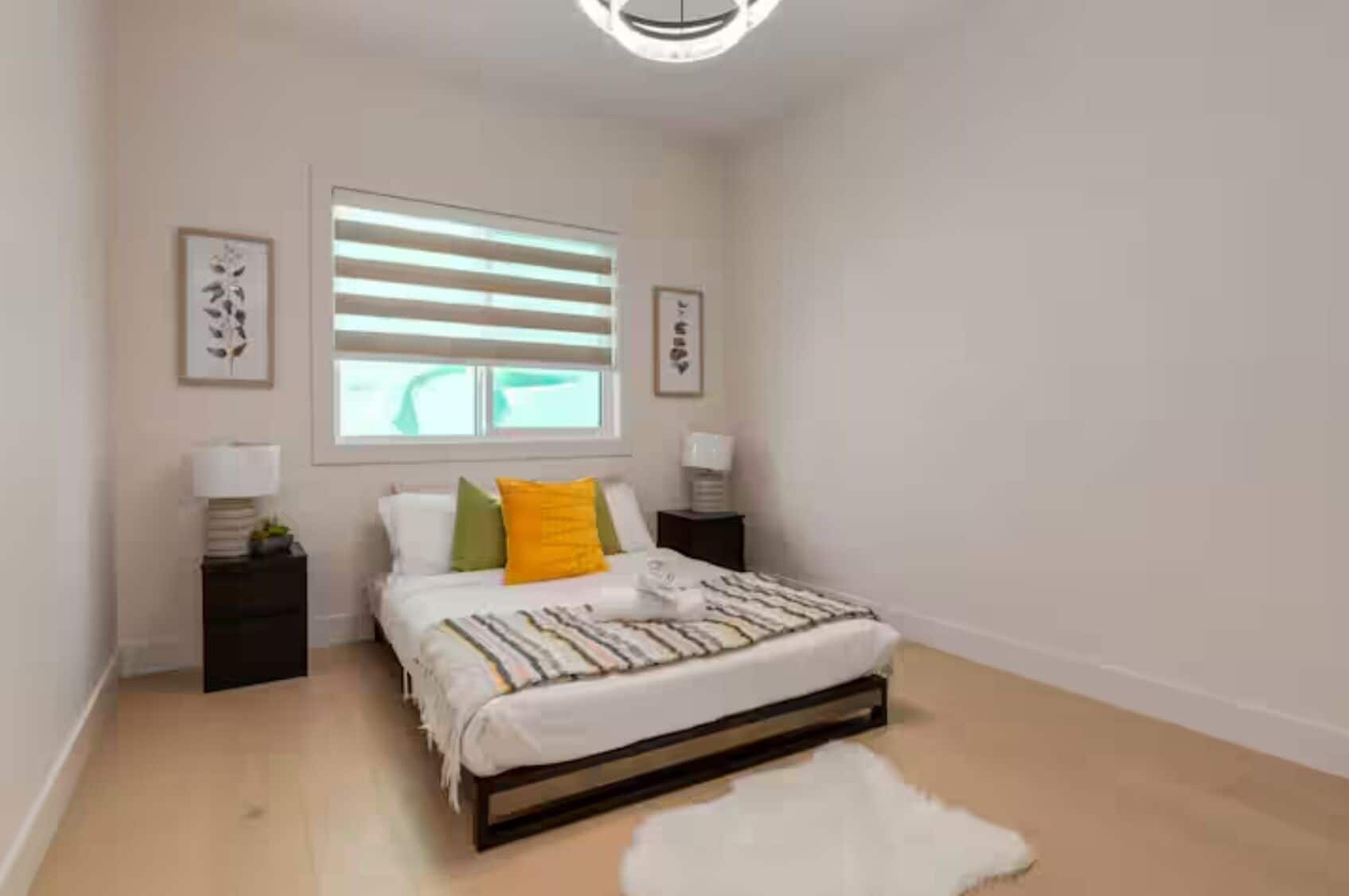 A bedroom with a bed, bedside tables, a window with blinds, and framed artwork on the walls.