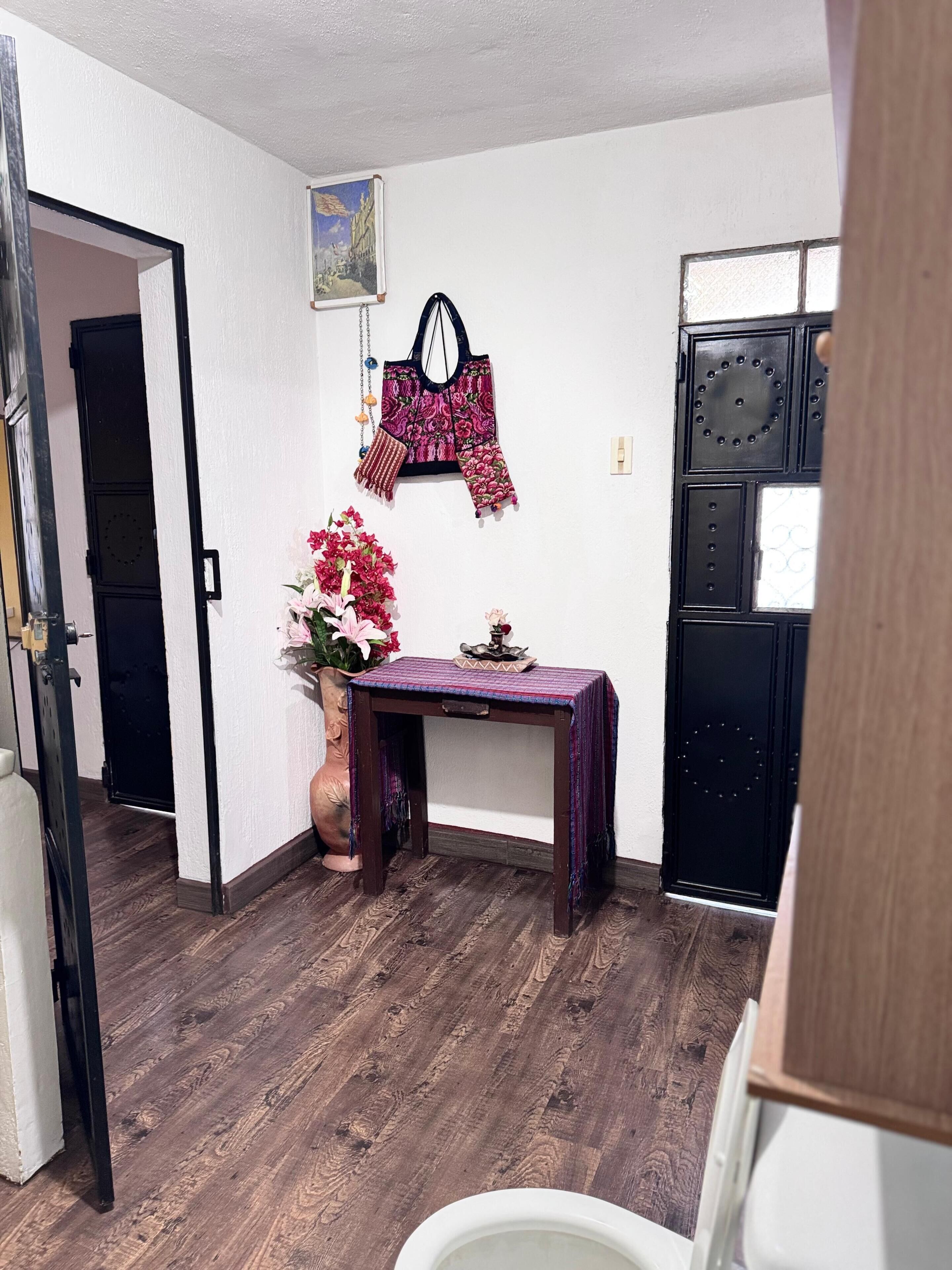 A room with a wooden floor, a small table with a purple cloth, a potted plant, and a black refrigerator.