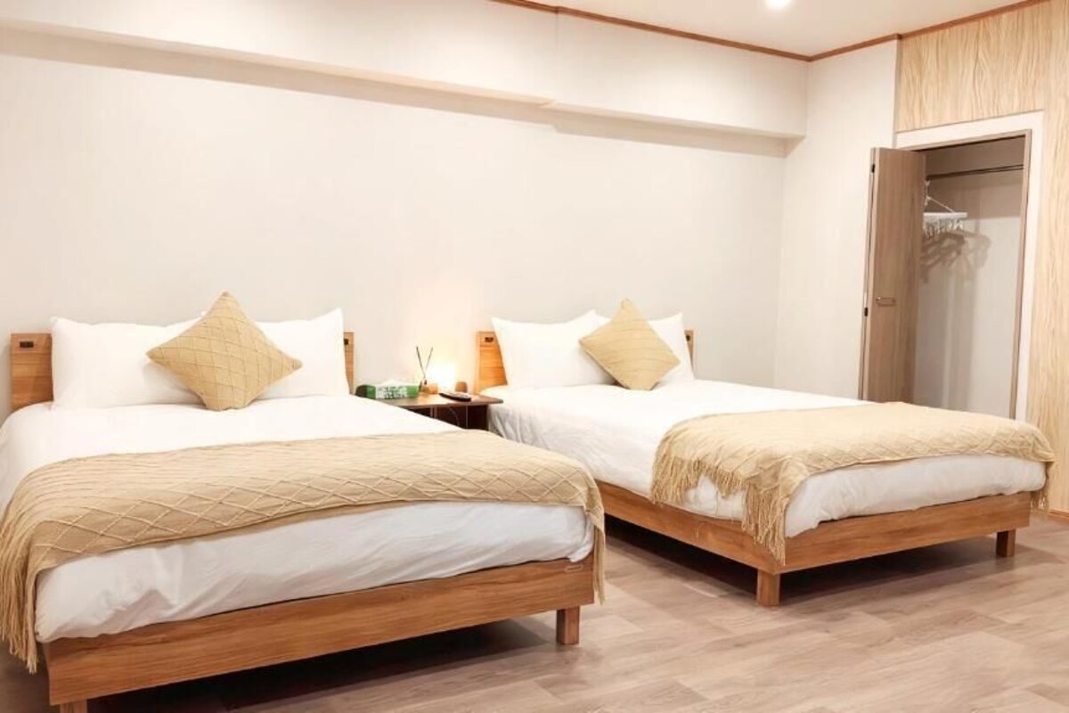Due letti singoli con struttura in legno, biancheria bianca e cuscini decorativi beige in una stanza con pavimento in legno e un armadio con porta in legno.