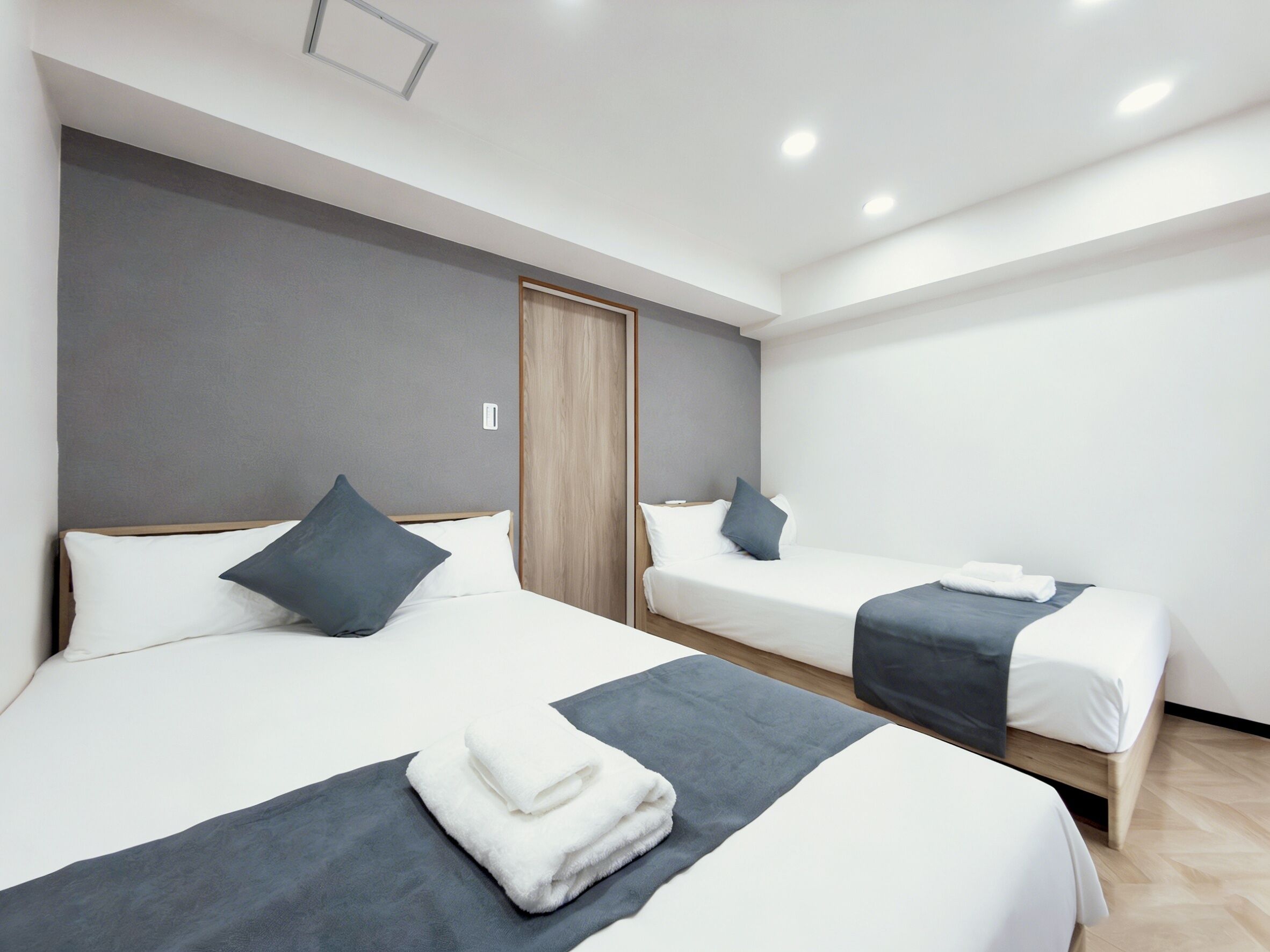 A modern hotel room with two beds, grey walls, wooden headboard, and a ceiling light.