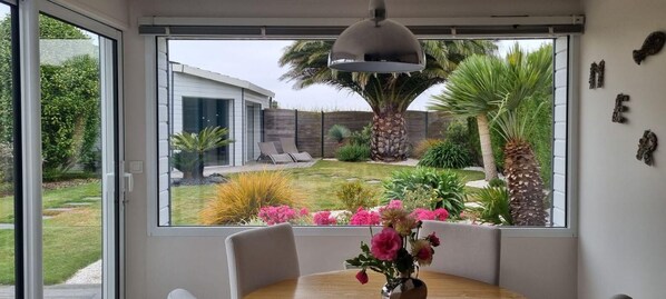 En matplats med ett bord och stolar, en vas med blommor och utsikt över ett modernt hus och en trädgård genom stora fönster.