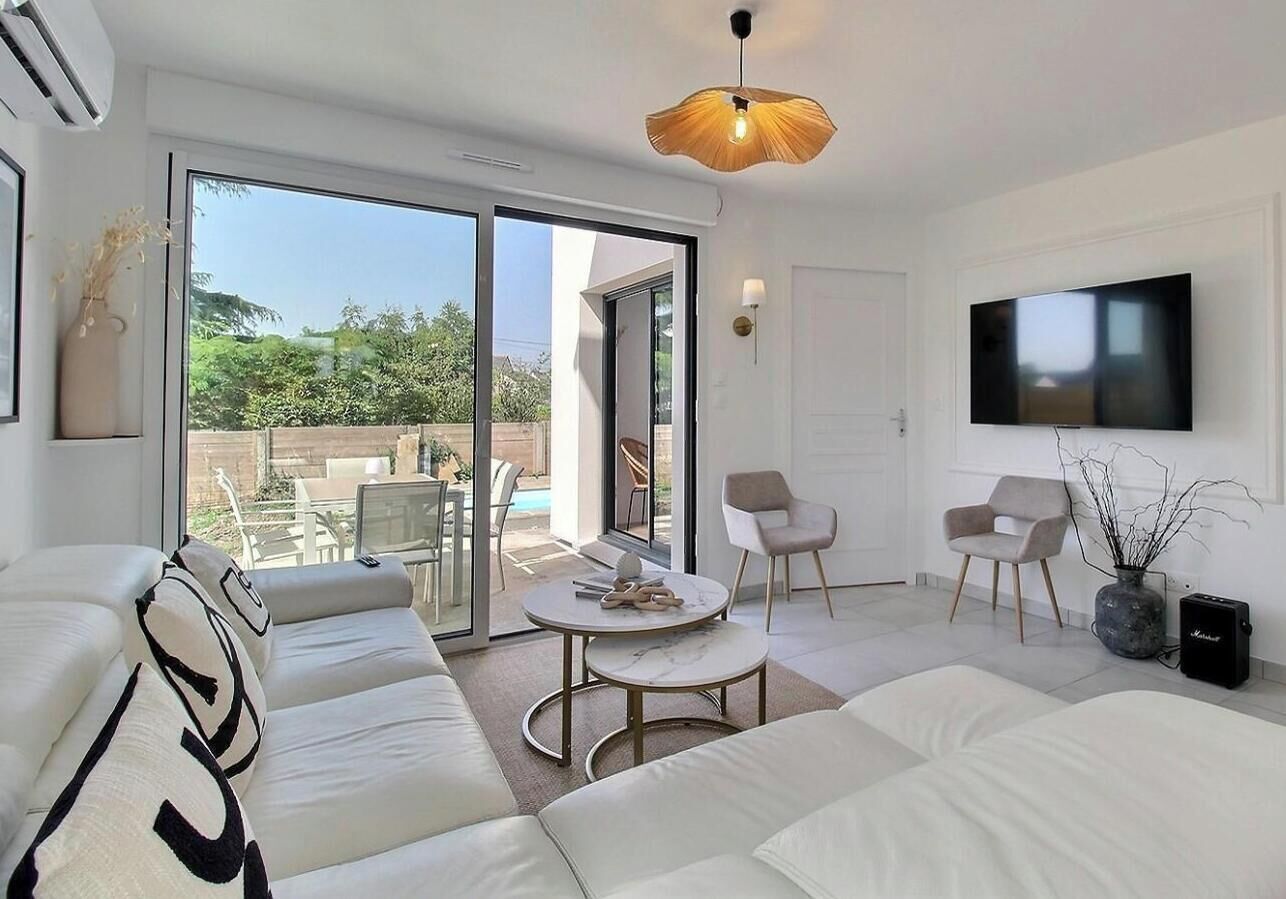 A modern living room with a sofa, a coffee table, and a TV. There is a sliding glass door leading to an outdoor area with a pool.