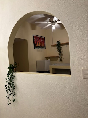 A room with a white textured wall, a ceiling fan, a small refrigerator, a plant, and a framed artwork. - Del Sol Suite: Ideal for travelers who enjoy a quiet place to relax (Puerto Vallarta)