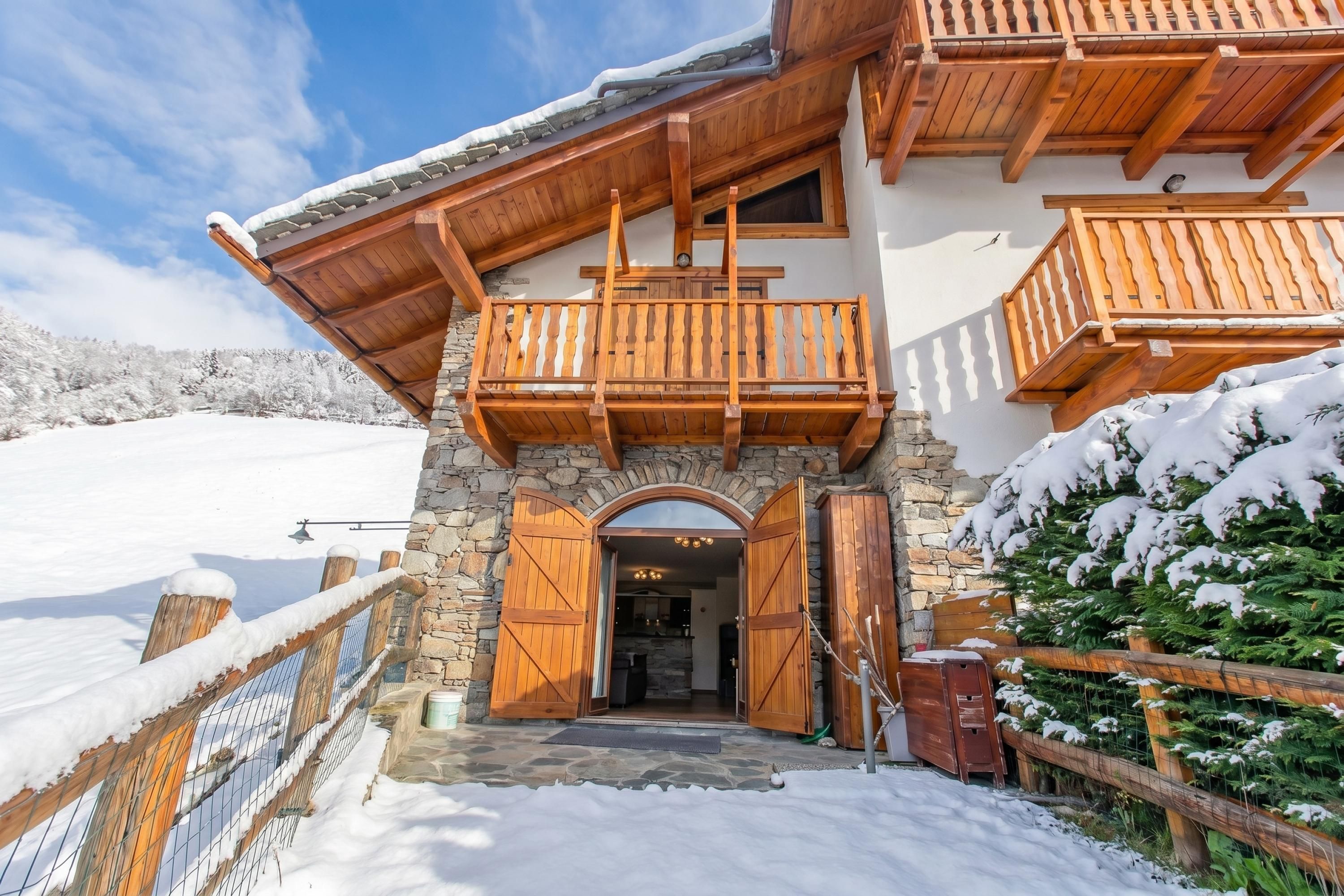 Un chalet à la façade en pierre, doté de balcons en bois et dont l’entrée est recouverte de neige.