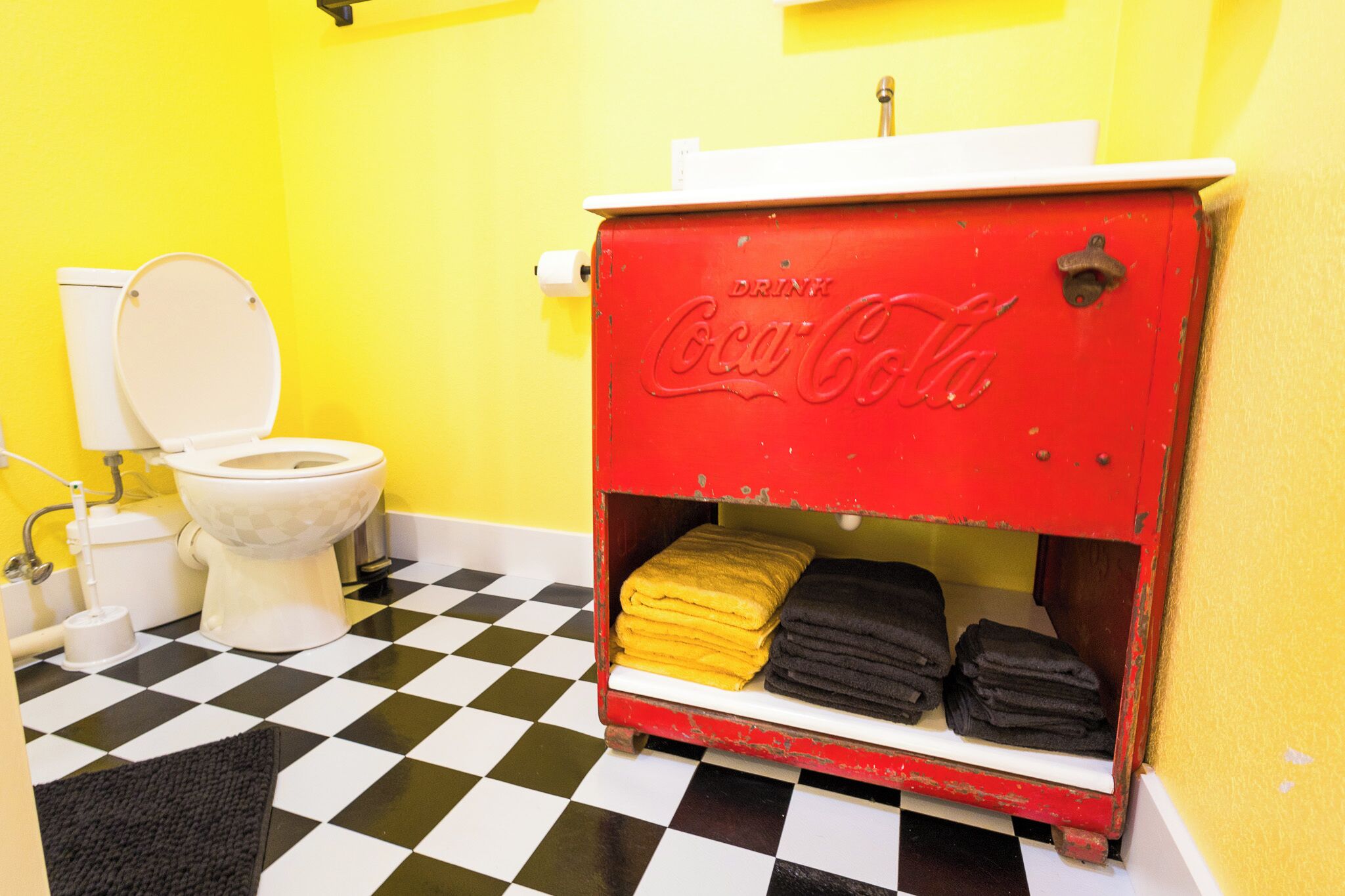 A bathroom with a red Coca-Cola vending machine, a toilet, and a checkered floor.