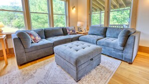 A living room with a sectional sofa, ottoman, and large windows offering a view of greenery.
