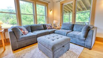 A living room with a sectional sofa, ottoman, and large windows offering a view of greenery.