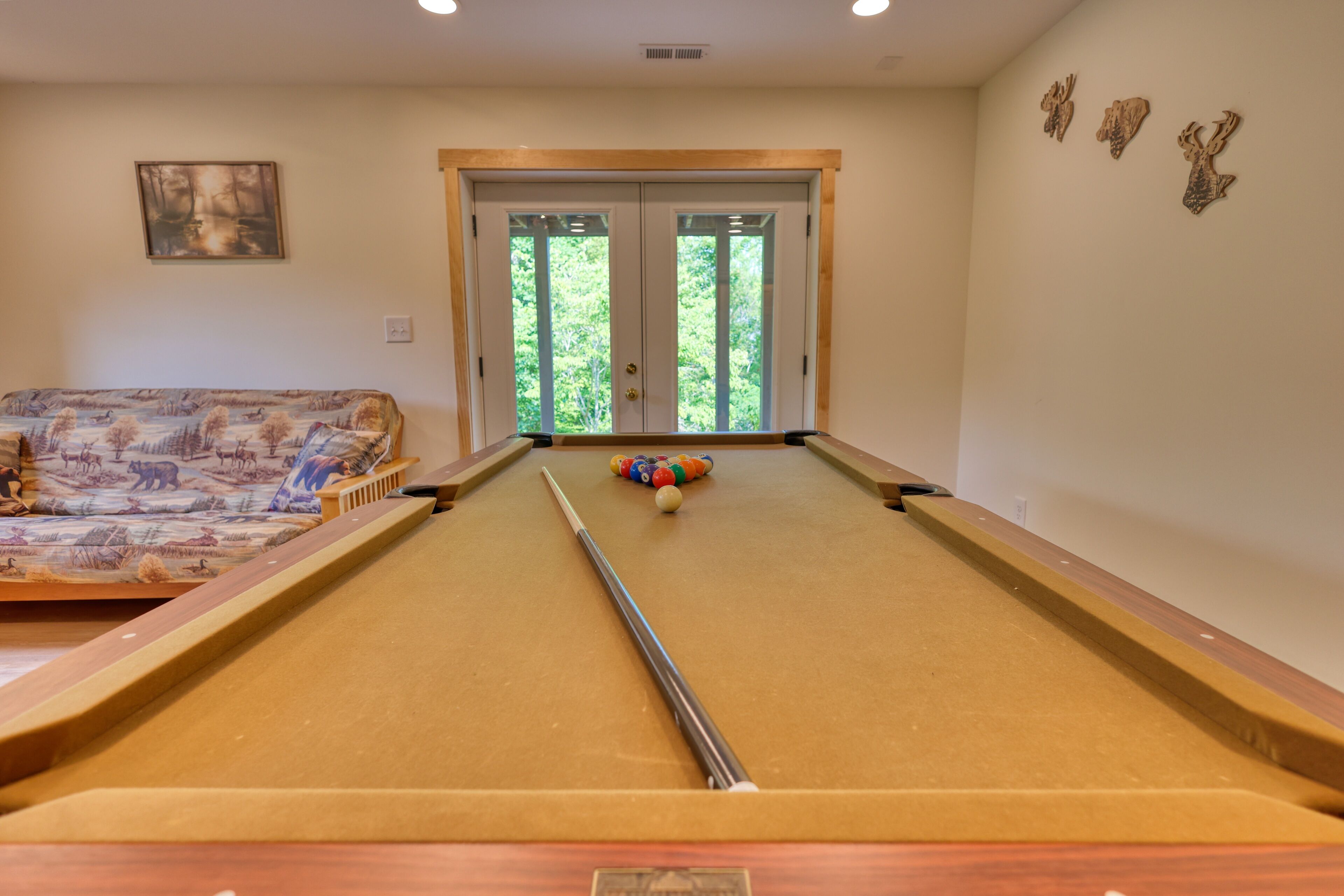 A pool table with a pool cue, a sofa with a forest print, and a door leading to a green outdoor area.