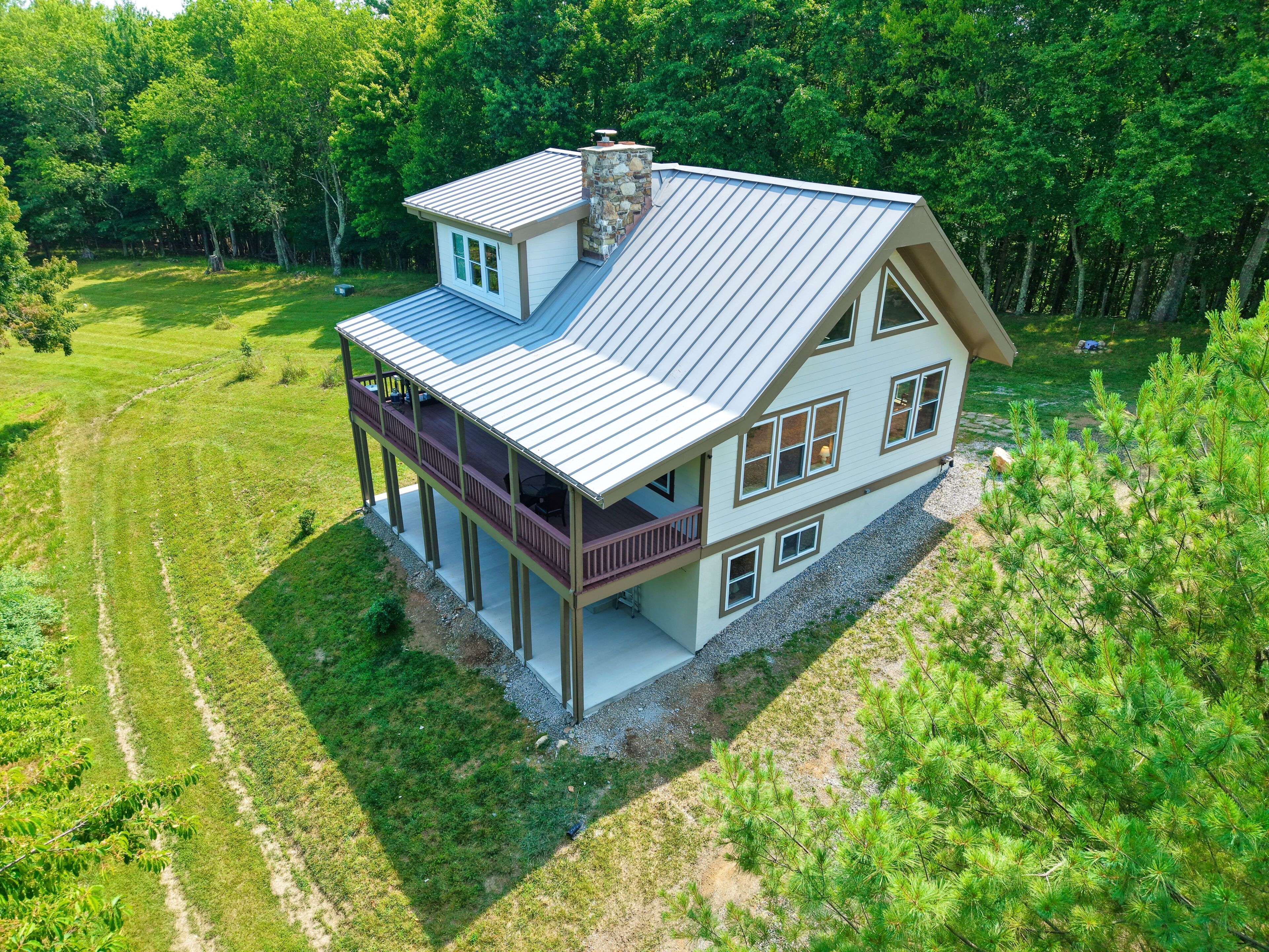 Une maison à deux étages dotée d’un balcon, d’une cheminée en pierre et entourée d’une zone boisée.