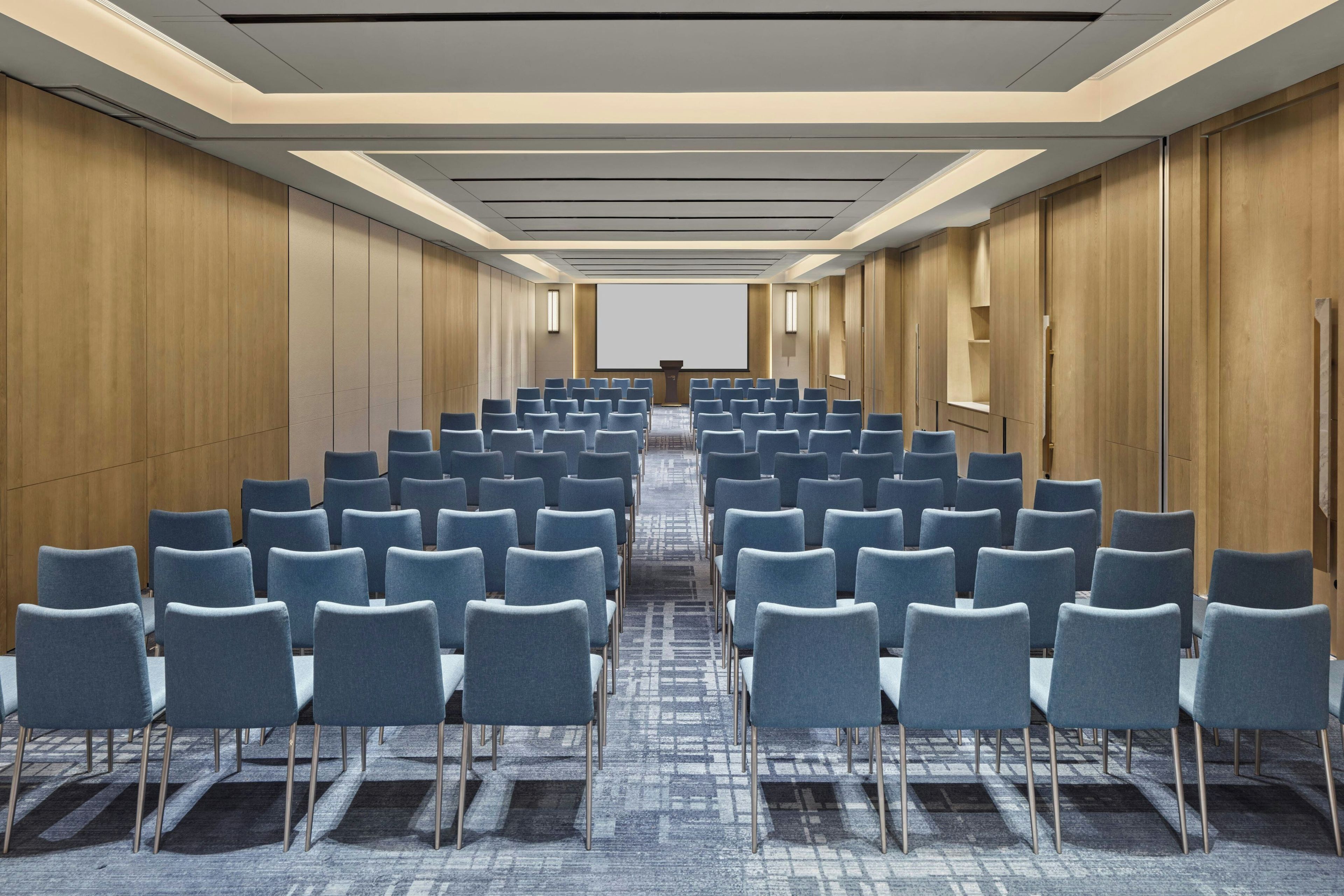 Sala de conferências com fileiras de cadeiras, uma tela de projeção e painéis de madeira nas paredes.