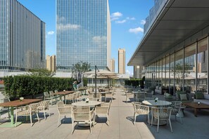 Un área de estar al aire libre con mesas y sillas, rodeada por un seto y con vista a un paisaje urbano con edificios altos.