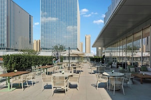 An outdoor seating area with tables and chairs, surrounded by a hedge and overlooking a cityscape with tall buildings.