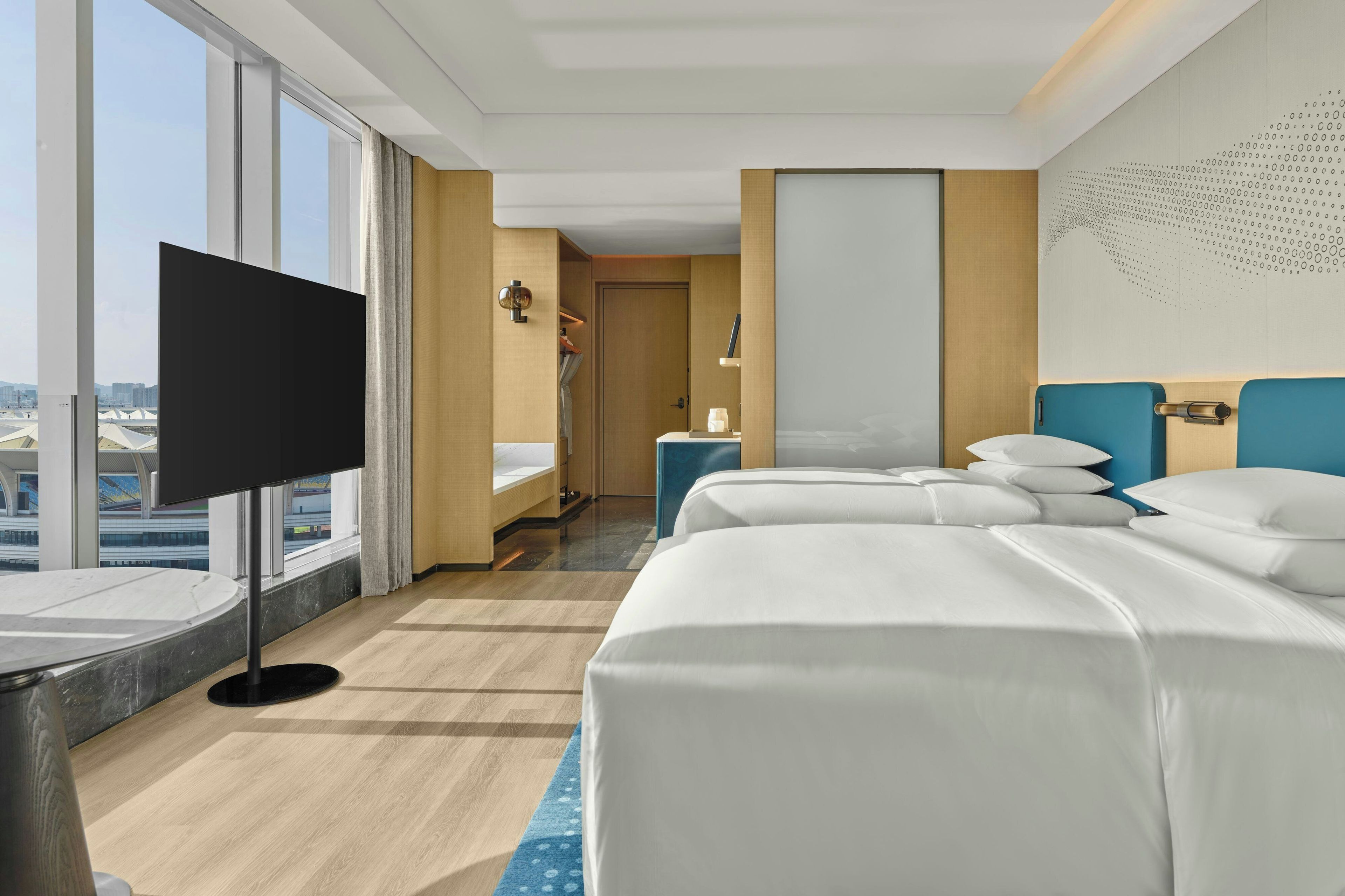 A hotel room with a large window, a flat-screen TV, a bed with white linens, a bedside table, and a nightstand with a lamp.