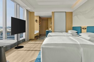 Une chambre d’hôtel dotée d’une grande fenêtre, d’un téléviseur à écran plat, d’un lit avec du linge de maison blanc, d’une table de chevet et d’une petite table de nuit avec une lampe.