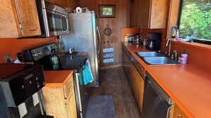 A compact kitchen with wooden cabinets, stainless steel appliances, a coffee maker, and a window with a view of greenery. - Charming 2-bedroom country house in Fort Bragg with a view of Mckerriker Beach. (Fort Bragg)