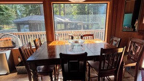 A wooden dining room with a large table and chairs, a chandelier, and a view of a deck and trees. - Charming 2-bedroom country house in Fort Bragg with a view of Mckerriker Beach. (Fort Bragg)