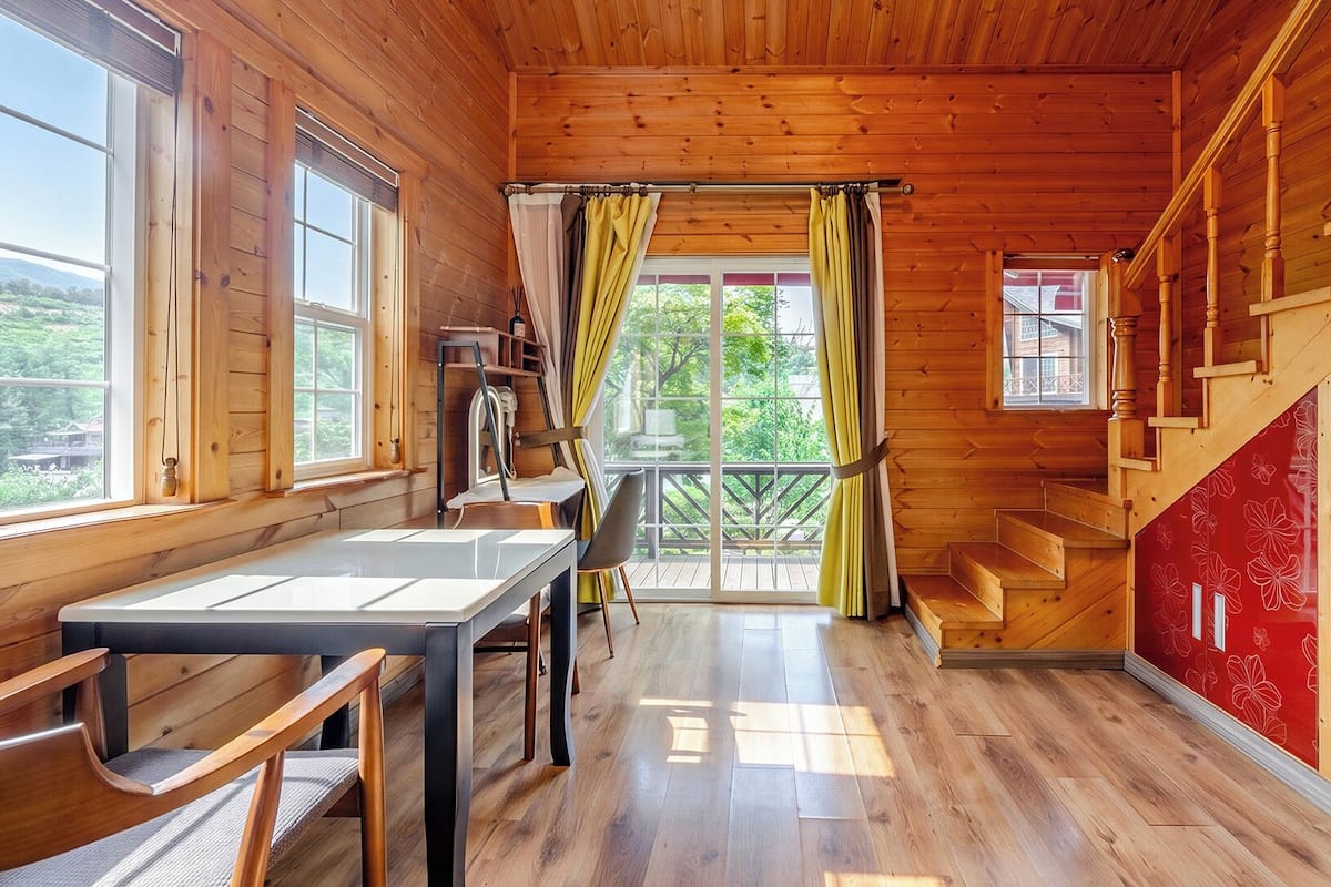 Interior de una cabaña de madera con una mesa de comedor, sillas, una escalera y vistas a través de un ventanal grande.