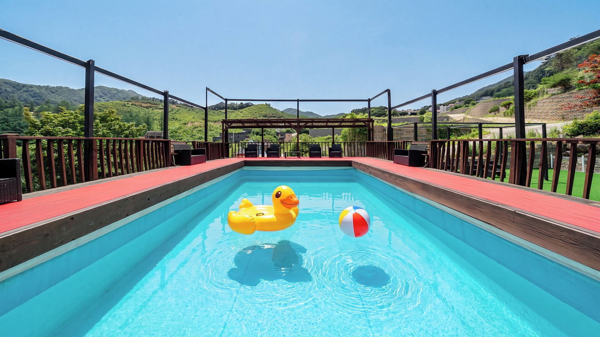 A swimming pool with a wooden deck, surrounded by a metal railing, and a view of hills in the background.