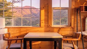 A wooden interior with a table and chairs, large windows with a mountain view, and a ladder against the wall.