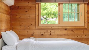 A wooden cabin room with a bed, a window with a view of greenery, and a wall-mounted air vent.