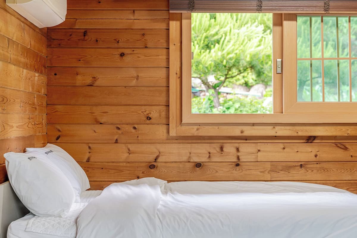 Una habitación de cabaña de madera con cama, ventana con vistas a follaje y un respiradero en la pared.