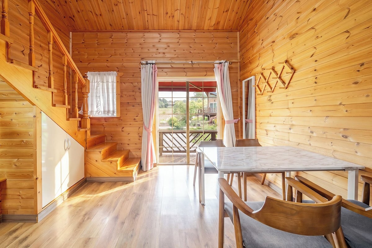 Interior de una cabaña de madera con comedor, escalera y vistas al exterior por la ventana.
