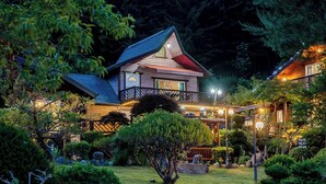 A two-story wooden house with a sloped roof, a balcony, and a well-lit garden.