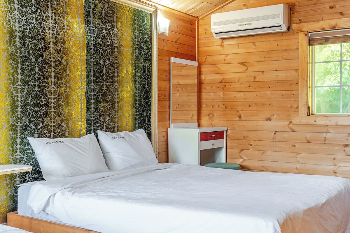 Un dormitorio de cabaña de madera con cama, mesita de noche, cómoda y un equipo de aire acondicionado, además de una ventana con vistas a la vegetación.