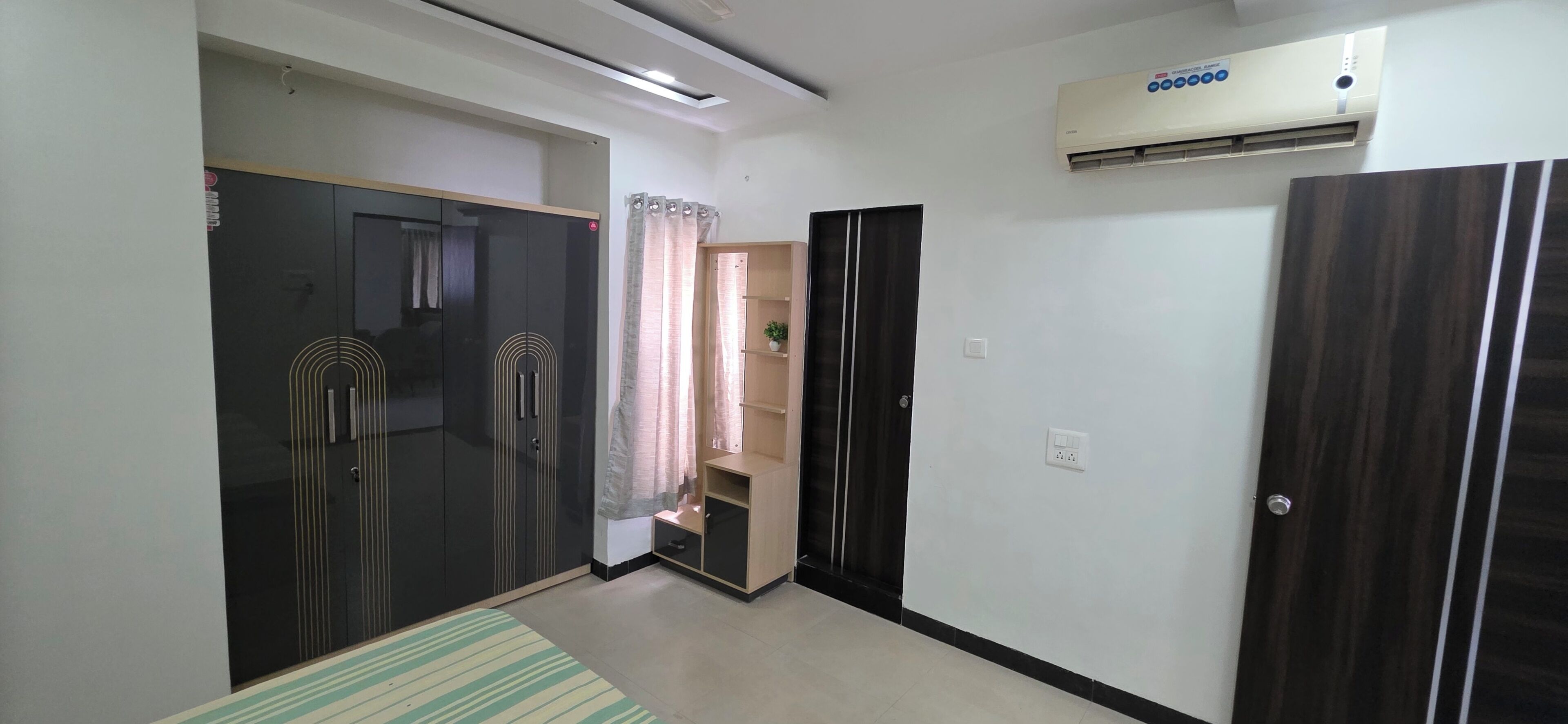 A room with a wardrobe, a small shelf with a plant, a door, and an air conditioning unit.