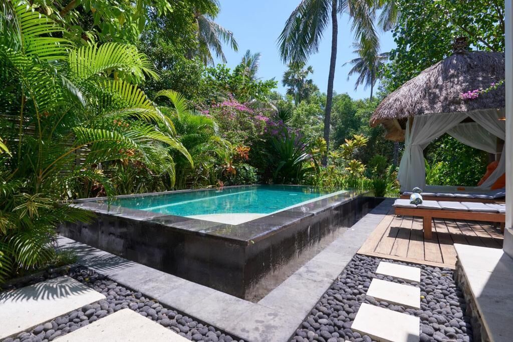 A rectangular pool surrounded by tropical vegetation, a wooden deck, and a thatched-roof structure.