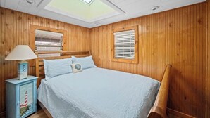 A bedroom with a skylight, wooden walls, a bed with a white bedspread, a nightstand with a lamp, and a window with blinds. - Dockside Cozy Cottage/Boat access/Spacious Yard/ sleeps 8 (Port Clinton)
