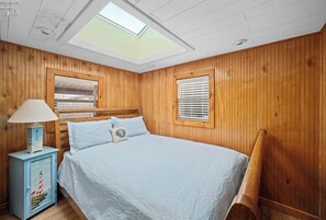 A bedroom with a skylight, wooden walls, a bed with a white bedspread, a nightstand with a lamp, and a window with blinds. - Dockside Cozy Cottage/Boat access/Spacious Yard/ sleeps 8 (Port Clinton)