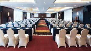 Un salón de conferencias con mesas y sillas en filas, suelo alfombrado e iluminación en el techo.
