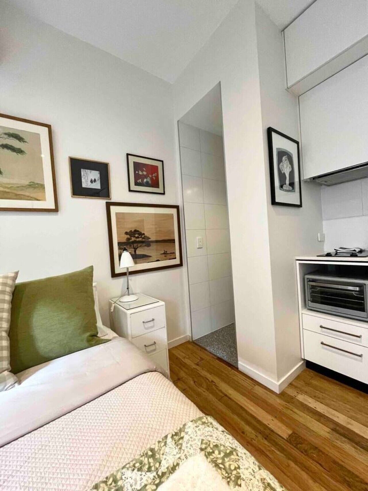 A bedroom with a bed, bedside table, lamp, and a small kitchenette with a microwave.