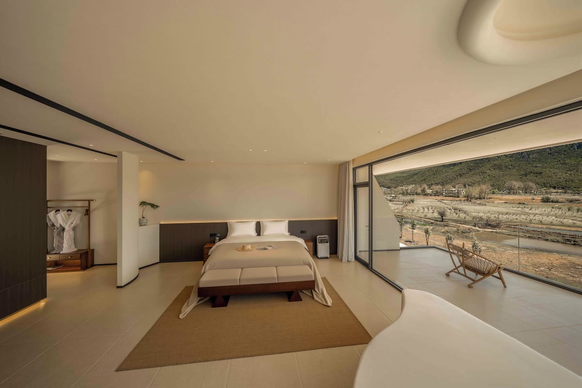 A modern bedroom with a large bed, a bedside table, a nightstand, a chair, and a view of a landscape through large windows.