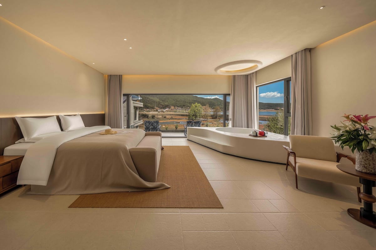 A modern hotel room with a large bed, a sofa, a view of a lake, and a balcony.