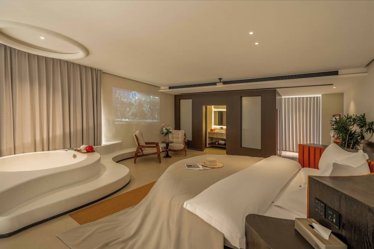 A modern hotel room with a large bathtub, a bed, a desk, and a seating area with chairs.