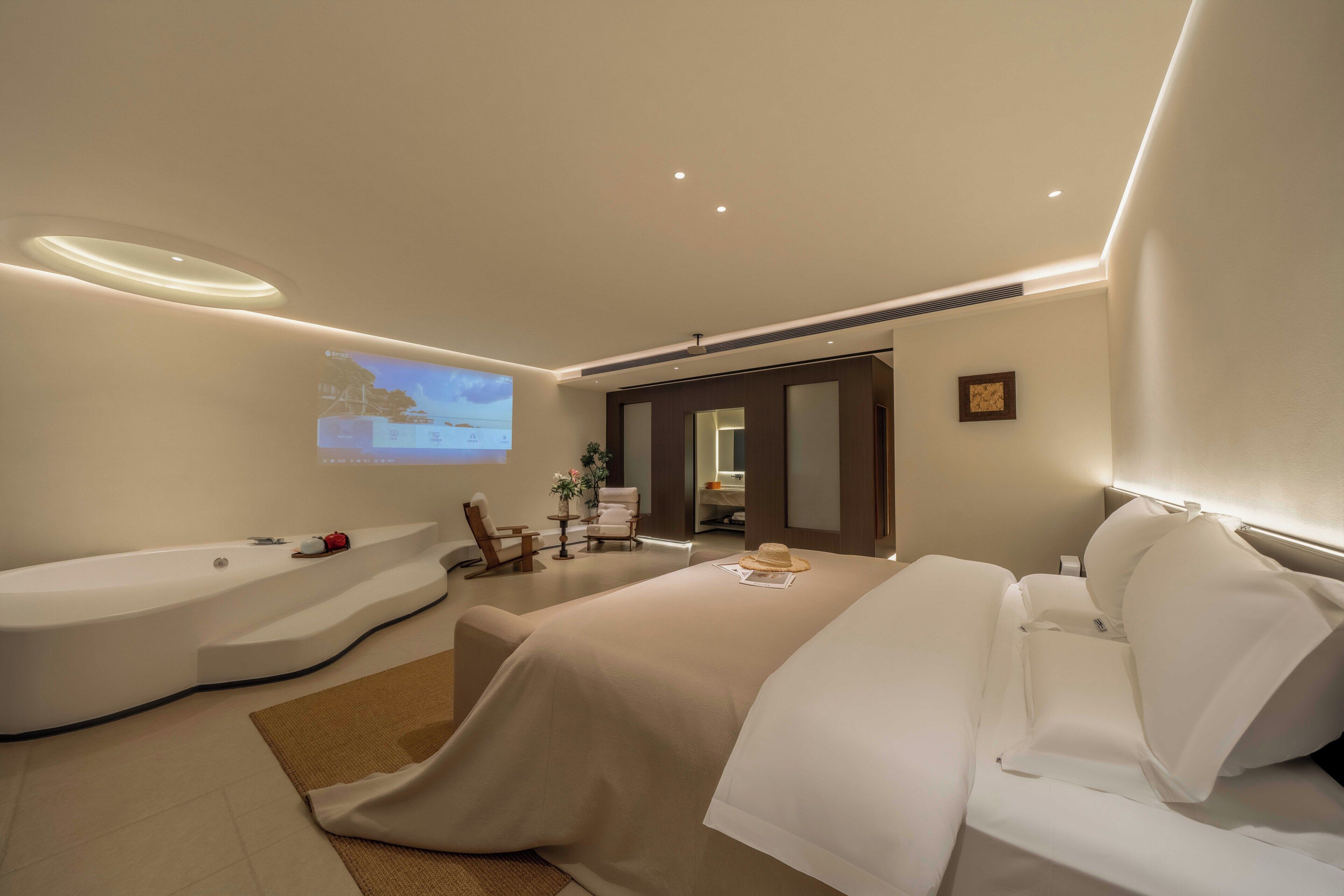 A modern hotel room with a large bed, a separate sitting area, a bathtub, and a projection screen on the wall.