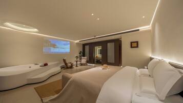 A modern hotel room with a large bed, a separate sitting area, a bathtub, and a projection screen on the wall.