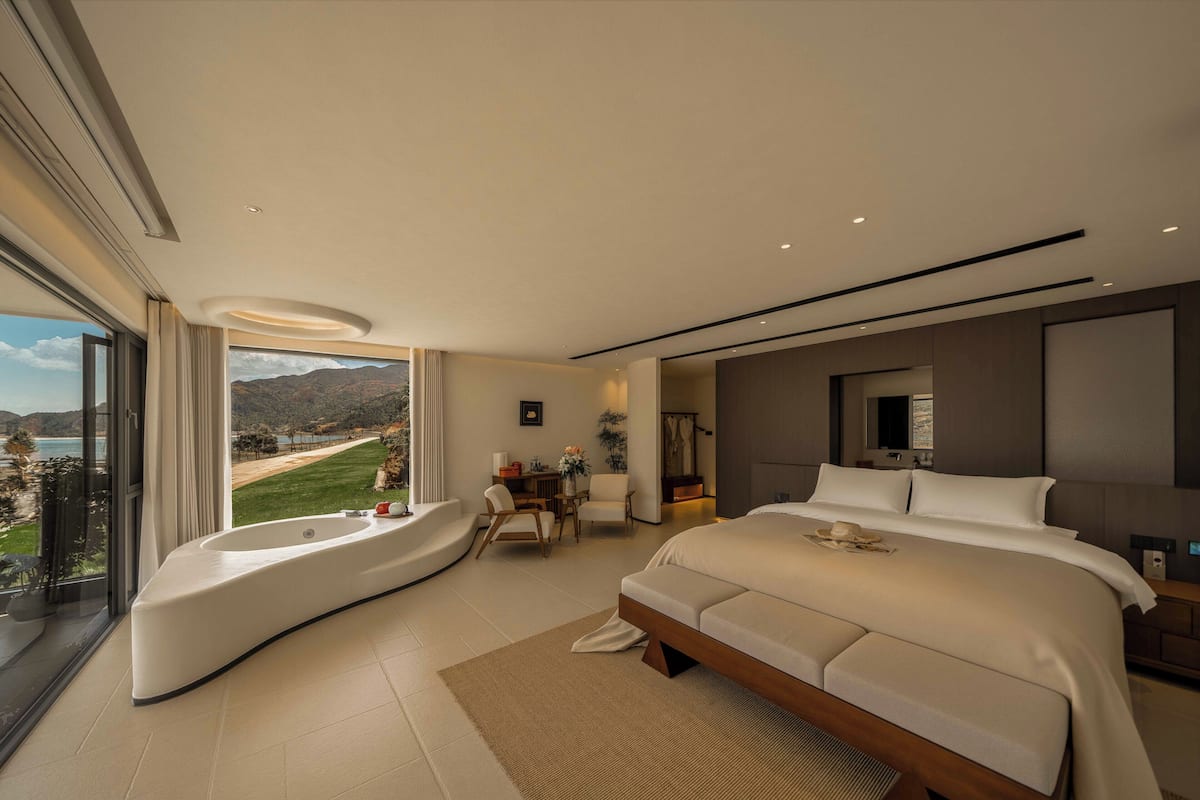 A modern hotel room with a large bed, a curved seating area, a desk, and a view of the outdoors.