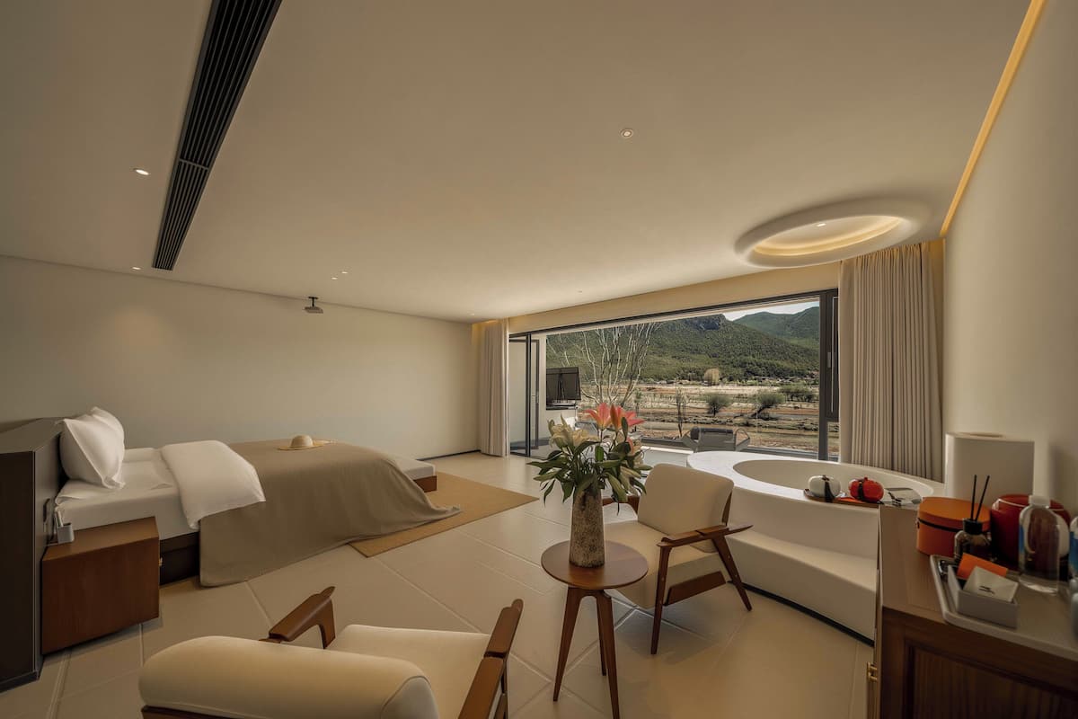 A modern hotel room with a bed, seating area, and a large window offering a view of the outdoors.