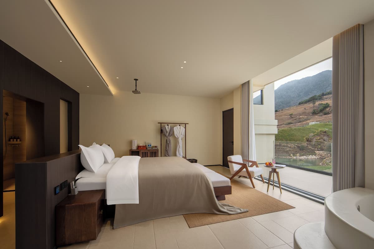 A modern hotel room with a large bed, a sitting area, a bathtub, and a view of the outdoors.